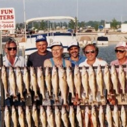 Group of smiling fishermen posing on a dock in front of a boat, holding a long row of freshly caught fish